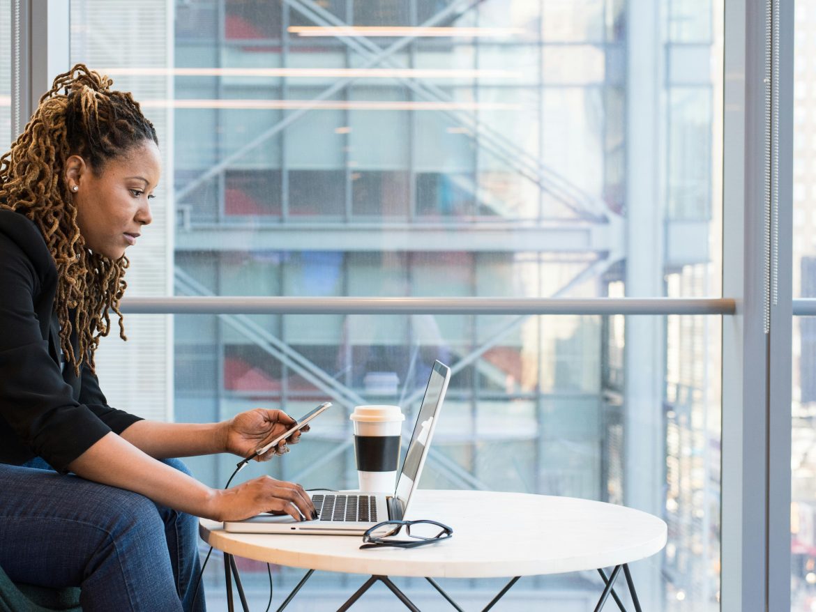 Woman working on a laptop and phone beside a large office window